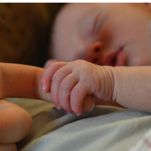 Zbliżenie na malutką dłoń noworodka chwytającą palec wskazujący dorosłego w odruchu chwytnym. Na nadgarstku dziecka widać szpitalną opaskę identyfikacyjną. Tekstura delikatnej skóry dziecka jest szczegółowa. Ciepłe, naturalne oświetlenie.