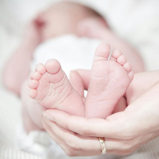 Zbliżenie na malutkie, pomarszczone stopy noworodka delikatnie trzymane w dłoniach dorosłego, na którego palcu widać złotą obrączkę. Na jednej kostce dziecka znajduje się szpitalna opaska identyfikacyjna. Tło to miękko rozmyte szpitalne pościeli. Naturalne, ciepłe światło.