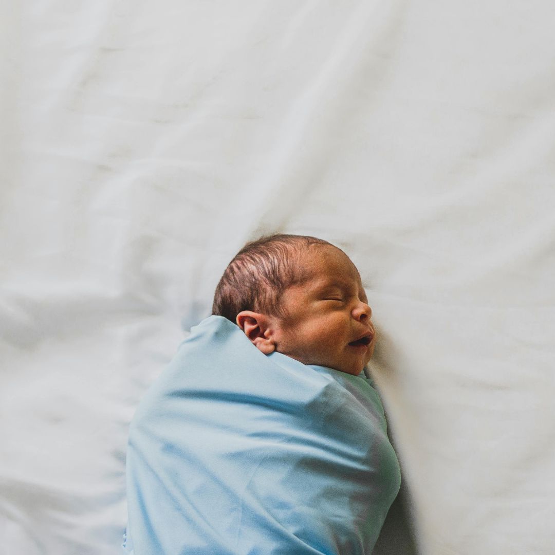 Śpiący noworodek owinięty ciasno w błękitny kocyk (rożek), leżący na białej pościeli. Widoczny profil twarzy dziecka i ciemne włoski.